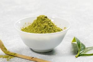 tazón blanco de primer plano lleno de polvo de té verde matcha, cuchara especial de bambú y hojas frescas sobre fondo gris texturizado. foto
