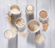 Different glass containers with oat milk, bowls with oat seeds and flakes on white wooden background. photo