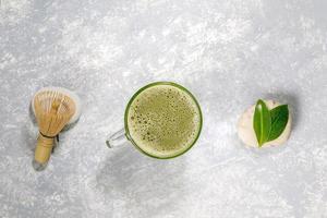 Glass cup with japanese matcha green tea, bamboo whisk and leaves on stones laid out in row on grey textured backdrop. photo