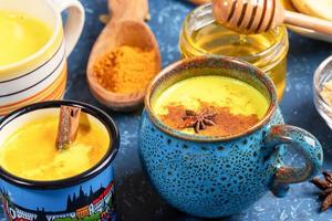Different cups with golden turmeric milk, curcuma powder and honey on blue textured backdrop. Close-up, selective focus. photo
