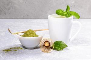 White cup with matcha green tea, powder with special wooden spoon, bamboo whisk, fresh leaves on grey backdrop. photo