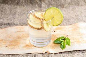 vaso con agua de jengibre y rodaja de limón y menta en un trozo de corteza sobre arpillera. foto