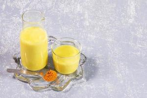 leche de cúrcuma orgánica. botella y vaso con leche dorada sobre placa transparente figurada sobre fondo gris claro. foto