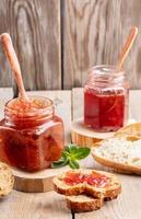 Two glass jars of strawberry and apple jam and sliced bread on wooden background. photo