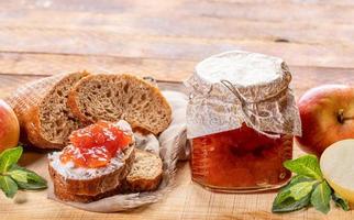 Small glass jar with apple jam, bread, smeared with apple jam, and apples and  mint on wooden background. photo