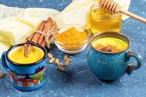 Golden turmeric milk in two blue cups and ingredients for its cooking on yellow napkin. Blue backdrop. Selective focus. photo