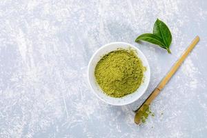 polvo de té matcha verde asiático en tazón blanco, cuchara especial de bambú de madera y hojas frescas sobre fondo gris texturizado. foto