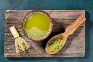 Japanese green matcha tea in wooden cup, matcha powder in wooden spoon and bamboo whisk on old wooden board. Top view. photo