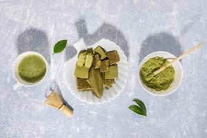 Matcha green tea, Matcha powder, treats containing matcha, whisk and leaves on light grey background with shadows. photo