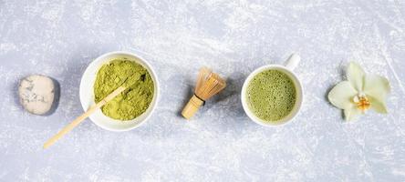 Cups with matcha green tea and powder, bamboo whisk, stone and orchid flower laid out in row on grey textured backdrop. photo