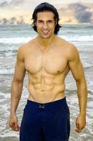 Handsome young man is in his swim suit at the beach at sunset showing off his incredibly fit body. photo