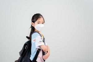 niña asiática lleva una mochila con máscara de respirador para proteger el brote de coronavirus y apunta con la mano al fondo en blanco, nuevo virus covid-19 foto