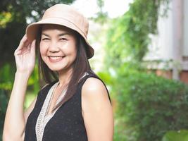Asian woman, wearing hat and black top sleeveless, standing in the garden, touching her hat,  smiling happily and looking at camera. photo