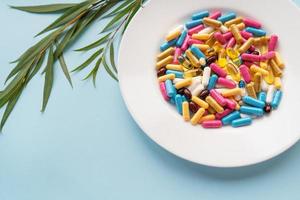 White plate with pills of nutritional supplements in different bright colors. Blue background, place for an inscription. Green branch on the background. photo