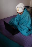 Home office, telecommuting, isolation, quarantine. A young girl working on a laptop, sitting on a bed at home in a towel after a shower. photo