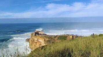 les gens regardent les grandes vagues géantes se briser près du phare du fort de nazare à nazare, au portugal. plus grosses vagues du monde. destination touristique pour le surf. video