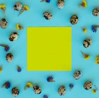 Easter composition. Quail eggs, flowers, yellow paper blank on blue background. Copy space. photo