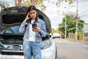 Angry Asian woman and using mobile phone calling for assistance after a car breakdown on street. Concept of vehicle engine problem or accident and emergency help from Professional mechanic photo