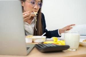 mujer de negocios ocupada y cansada comiendo espagueti para el almuerzo en la oficina y trabajando para entregar estados financieros a un jefe. con exceso de trabajo y poco saludable para comidas preparadas, concepto de agotamiento. foto