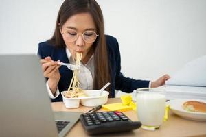 mujer de negocios ocupada y cansada comiendo espagueti para el almuerzo en la oficina y trabajando para entregar estados financieros a un jefe. con exceso de trabajo y poco saludable para comidas preparadas, concepto de agotamiento. foto