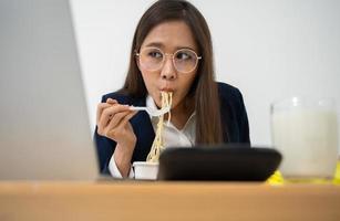 mujer de negocios ocupada y cansada comiendo espagueti para el almuerzo en la oficina y trabajando para entregar estados financieros a un jefe. con exceso de trabajo y poco saludable para comidas preparadas, concepto de agotamiento. foto