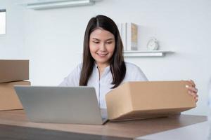 Happy young Asian woman entrepreneur, Hold package and Smile for sale success of online shopping store at home office, Concept of merchant, small business, online business and eCommerce. photo