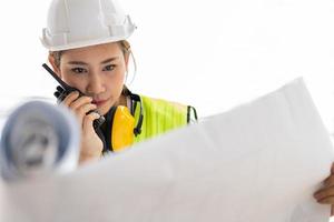 una ingeniera asiática o una joven arquitecta se ponen un casco por seguridad y miran el plano para inspeccionar el sitio de construcción de la fábrica y usan walkie talkie para hablar con un contratista.