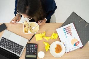 vista superior de una mujer de negocios ocupada y cansada comiendo espaguetis para almorzar en la oficina y trabajando para entregar estados financieros al jefe. con exceso de trabajo y poco saludable para comidas preparadas, concepto de agotamiento. foto