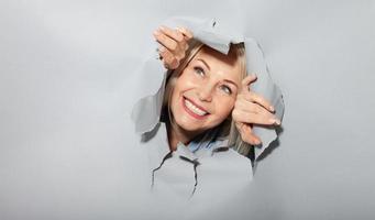 Surprised woman looking playfully in torn paper hole, has excited cheerful expression, looks through breakthrough of gray background. Wow, great news photo