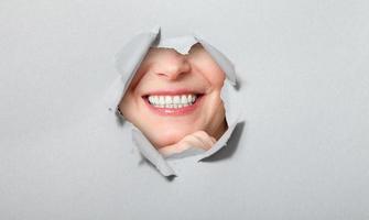 Surprised woman looking playfully in torn paper hole, has excited cheerful expression, looks through breakthrough of gray background. Wow, great news photo