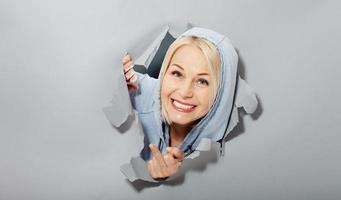 Charismatic joyful woman with blonde hairstyle, looking playfully, wears fashionable hoody, stands in torn paper hole, notices funny pleasant. Gray background. photo