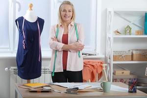 A successful woman designer clothes, tailor in the studio in full growth. Beautiful European woman looking at the camera, standing near the table in the workshop with clothes hanging in the background photo