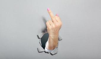 Woman hand shows with a gesture that everything middle finger . Torn hole in gray paper. photo