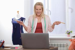 Communication with suppliers by phone, skype. Beautiful European woman tailor talking on the phone, standing near the table in the workshop with clothes hanging in background photo