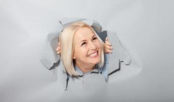 Charismatic joyful woman with blonde hairstyle, looking playfully, wears fashionable hoody, stands in torn paper hole, notices funny pleasant. Gray background. photo