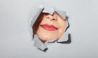 Woman showing her lips playfully in torn paper hole, through breakthrough of gray background. Cosmetics advertising concept photo