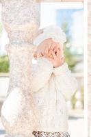 una niña rubia de pelo corto, con una diadema de flores, se cubre la cara con las manos. linda y pequeña niña en la calle. niña jugando al escondite foto