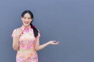 hermosa mujer asiática con el pelo largo que usa un vestido rosa cheongsam en el tema del año nuevo chino mientras su mano muestra algo y mira a la cámara sonriendo alegremente con el fondo gris. foto