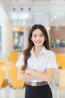 Portrait of an adult Thai student in university student uniform. Asian beautiful girl standing smiling happily and confidently with her arms crossed at the university reading room. photo