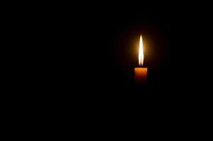 una sola llama de vela encendida o luz que brilla en una vela naranja sobre fondo negro u oscuro en la mesa de la iglesia para Navidad, funeral o servicio conmemorativo con espacio para copiar