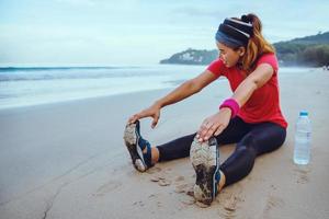 mujeres asiáticas jogging entrenamiento en la playa. sentarse en la playa fitness relajarse estirando las piernas y estirando el brazo. foto
