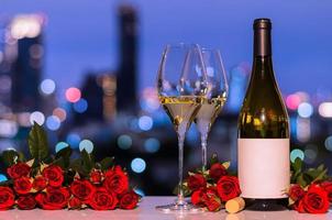 selectivo en la primera copa de vino blanco con botella borrosa y rosas rojas en la colorida luz del fondo de la ciudad para el concepto de cena de San Valentín. foto