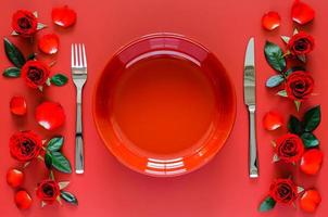 concepto de cena de san valentín con plato rojo, tenedor y cuchillo puesto en una mesa roja con rosas, pétalos y hojas. foto