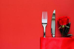 tenedor y cuchillo con rosa roja puesta en una servilleta sobre una mesa roja para el concepto de cena de san valentín. foto