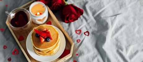 Pancakes in the shape of a heart with berries, roses flowers, cup of tea and candle in candlestick. Valentine's day breakfast concept. Top view with copy space. Banner for design, web site photo