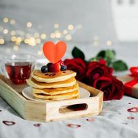 Valentine's day composition with breakfast, gift box and roses. Stack of delicious pancakes with berries and cup of hot moning tea on wooden tray photo