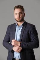 A handsome bearded man in a jacket and shirt on a gray background looks at the camera. photo