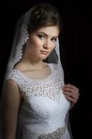 Beautiful bride with a wedding hairstyle - on a dark background. Portrait of a gorgeous bride. Wedding. The bride in a white dress holds a veil. photo