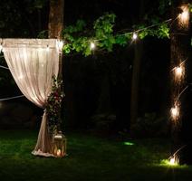 Wedding twilight ceremony decoration made of hanging on a tree golden lanterns and lamps and standing on the grass fired candles with the carpet in the middle photo