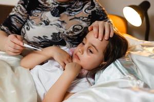 joven madre revisando la temperatura de la hija enferma acostada en la cama foto